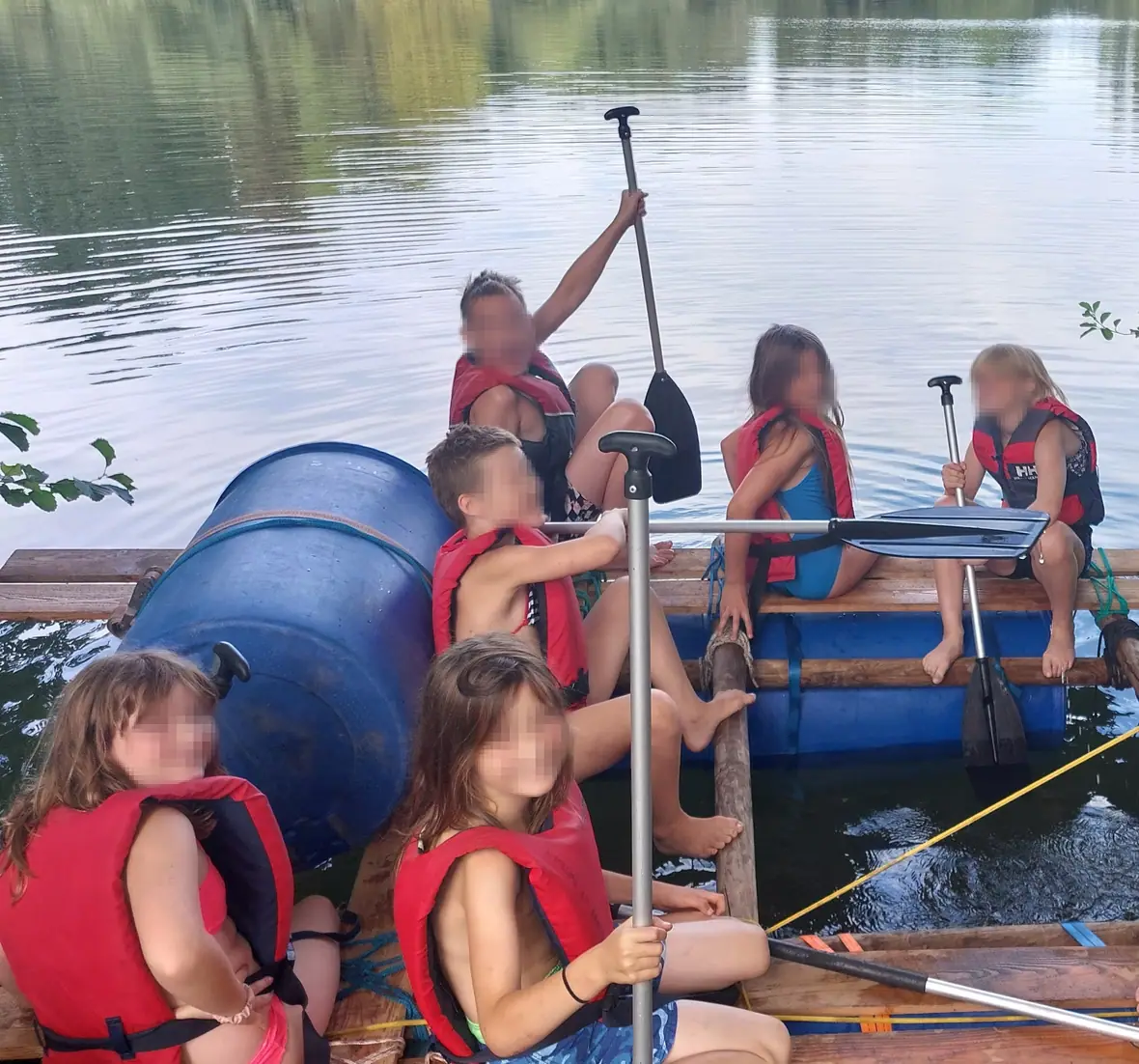 Eine Kindergruppe paddelt mit Schwimmwesten auf einem selbst gebauten Floß über einen See.