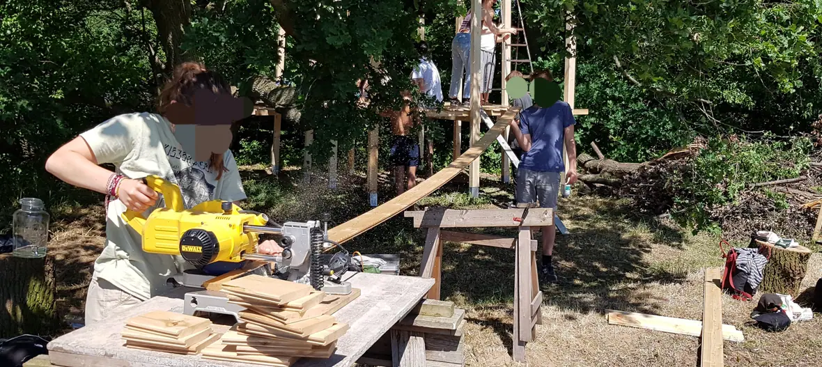 Jugendliche bauen gemeinsam mit Holz im Wald und sägen Bretter zu.