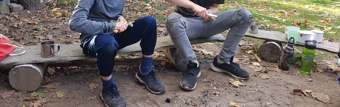Zwei Kinder schnitzen auf einer Bank im herbstlichen Wald an Stöcken.