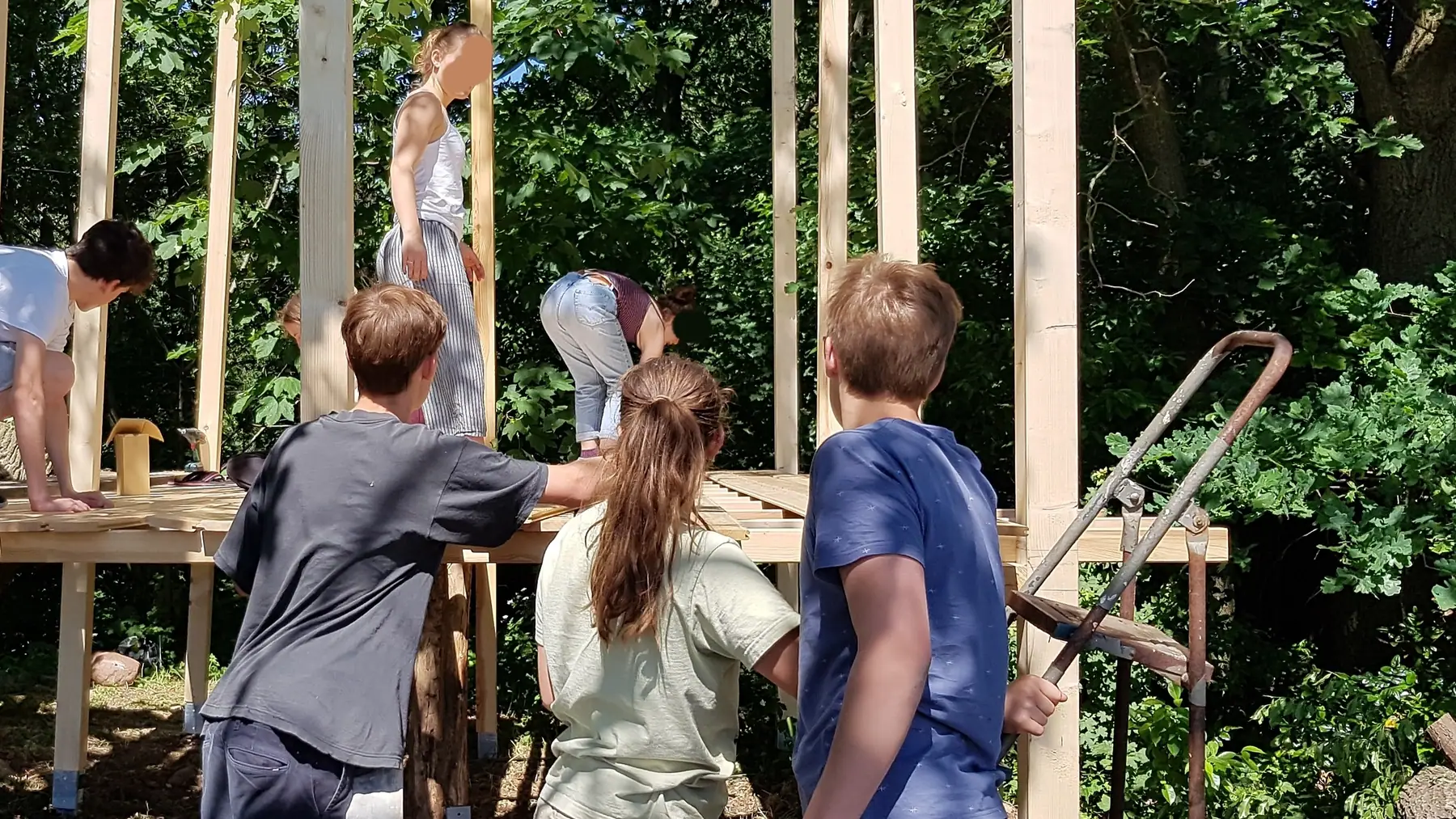 Kinder bauen gemeinsam ein Holzgerüst in einem Waldcamp, von hinten fotografiert.