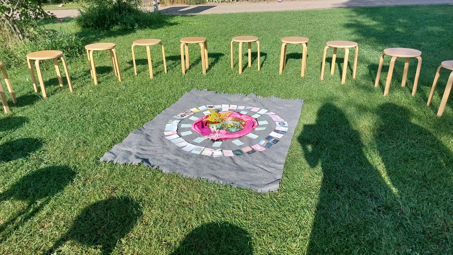 Ein Sitzkreis aus Holzhockern um ein Naturmandala mit Blumen auf einer sonnigen Wiese.