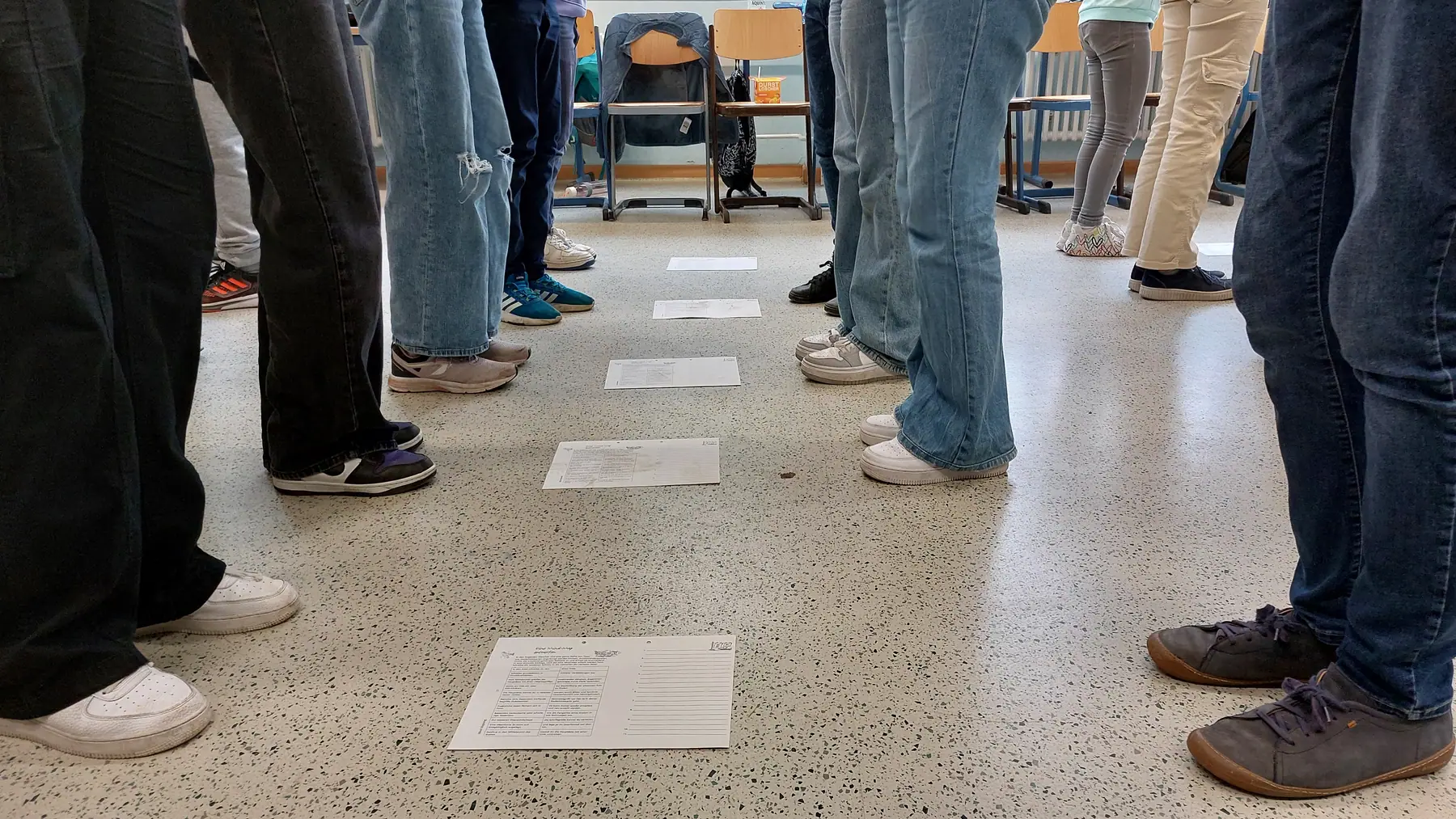 Eine Gruppe steht in einem Klassenraum um beschriftete Karten am Boden, nur Beine und Füße sichtbar.