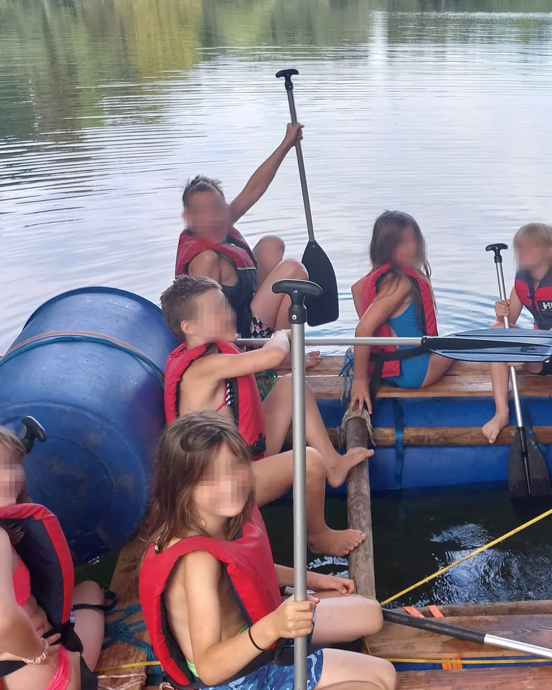 Eine Kindergruppe paddelt mit Schwimmwesten auf einem selbst gebauten Floß über einen See.